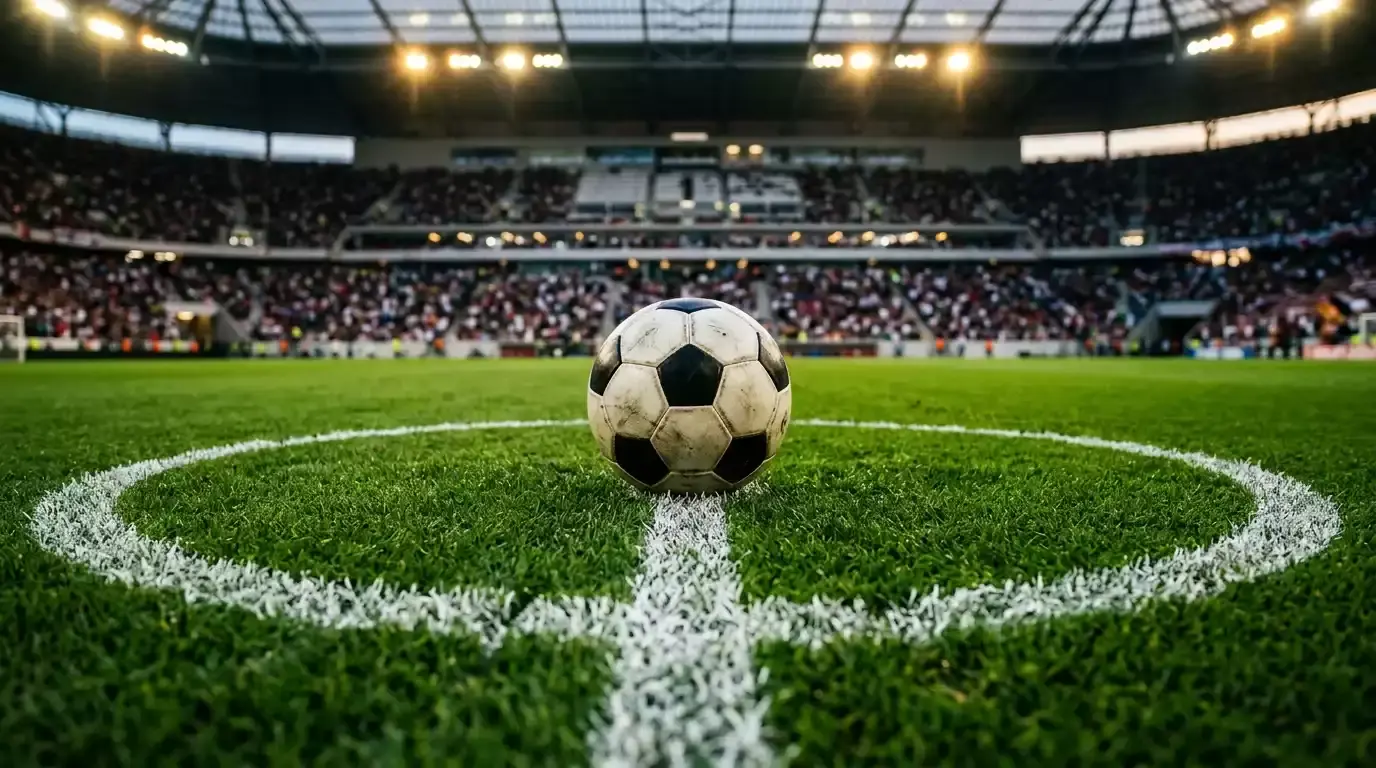 Futbalová lopta na trávniku pod večernými reflektormi, v pozadí rozostrený plný štadión, napätá derby atmosféra